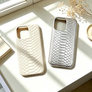 Könnte beinhalten: Zwei rechteckige Handyhüllen mit einer strukturierten Schlangenhaut-Optik. Eine Hülle ist in mattem Off-White gehalten, während die andere ein metallisch silbernes Finish aufweist. Beide haben einen quadratischen Ausschnitt für die Kamera.