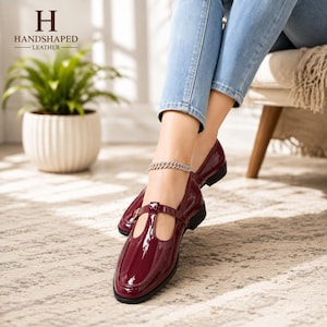 May include: A pair of dark burgundy patent leather T-strap shoes with a glossy finish, featuring a thin strap across the instep, a rounded toe, and a low block heel. The shoes are worn with light blue denim jeans and a silver chain anklet.