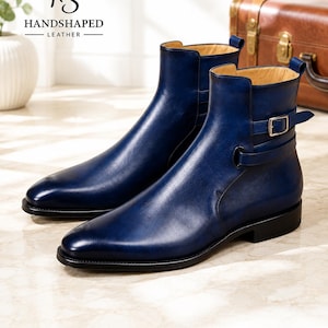 Könnte beinhalten: Ein Paar blaue Lederstiefeletten mit einem seitlichen Riemen mit silberfarbener Metallschnalle, einer glatten Oberfläche und einer schwarzen Sohle. Die Stiefel haben ein beiges Innenfutter und eine spitz zulaufende Form. Der Text HS HANDSHAPED LEATHER ist sichtbar.