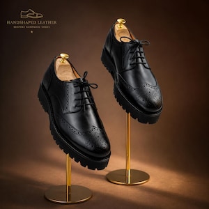 May include: Two black leather brogue shoes with thick soles are displayed on gold stands. The shoes have intricate perforations and lace-up closures. The text "HANDSHAPED LEATHER BESPOKE HANDMADE SHOES" is visible at the top.