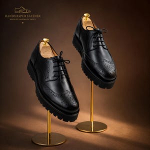 May include: Two black leather brogue shoes with thick soles are displayed on gold stands. The shoes have intricate perforations and lace-up closures. The text "HANDSHAPED LEATHER BESPOKE HANDMADE SHOES" is visible at the top.