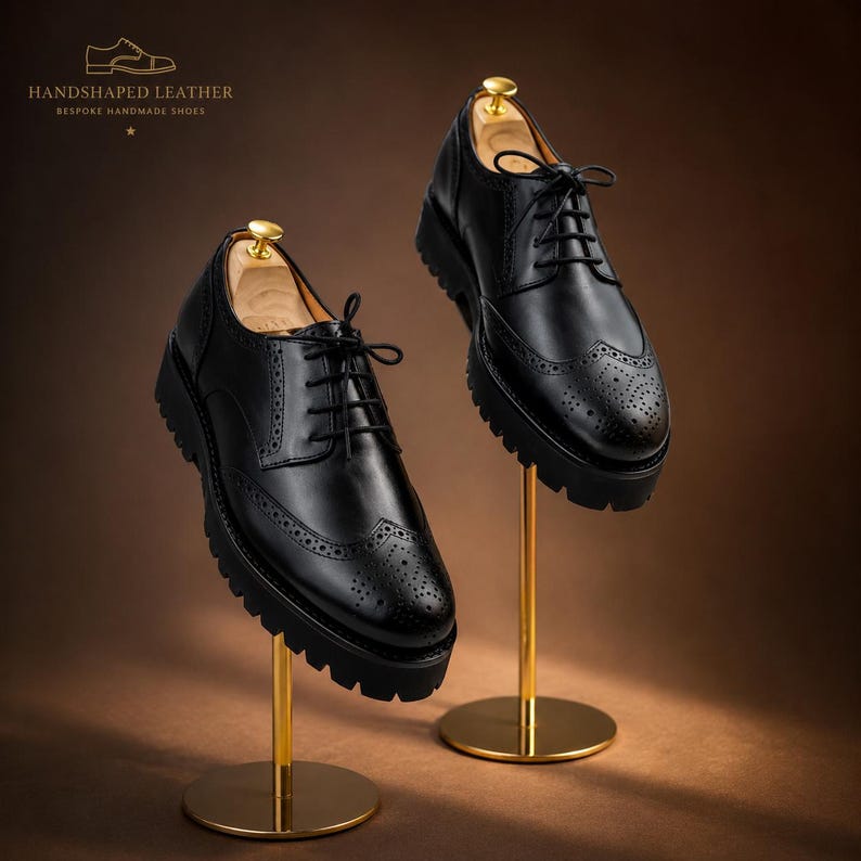 May include: Two black leather brogue shoes with thick soles are displayed on gold stands. The shoes have intricate perforations and lace-up closures. The text "HANDSHAPED LEATHER BESPOKE HANDMADE SHOES" is visible at the top.