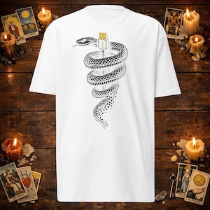 Op de afbeelding: Een wit t-shirt met korte mouwen en een zwart-witte afbeelding van een slang die om een gouden USB-connector is gewikkeld. De slang heeft een rode gespleten tong en het onderste deel van het lichaam gaat over in een digitaal pixelpatroon.