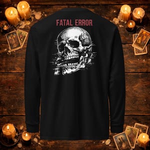 Puede incluir: Una camiseta negra de manga larga con un gráfico blanco de un cráneo humano con un efecto de distorsión digital en la mandíbula. El texto FATAL ERROR está impreso en color rojo sobre el cráneo.