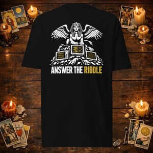 Op de afbeelding: Een zwart t-shirt met een witte afbeelding van een sfinx die op een stapel computermonitoren zit die binaire code en gegevens weergeven. Onder de afbeelding staat de tekst "ANSWER THE RIDDLE" in witte en gele hoofdletters.
