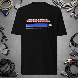 Op de afbeelding: Een zwart t-shirt met korte mouwen en een grafische print met de witte tekst "WEEKEND LOADING", een rode horizontale lijn, een blauwe voortgangsbalk op 99% en de kleine witte tekst "SYSTEM_STALL // ERROR_CODE: 0X99_FATAL".