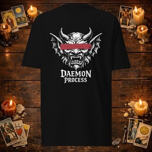 Op de afbeelding: Een zwart t-shirt met korte mouwen en een witte afbeelding van een gehoornde, gevleugelde demonenkop met een rode horizontale balk over de ogen. Onder de afbeelding staat de tekst DAEMON PROCESS in een wit, gestileerd schreeflettertype.