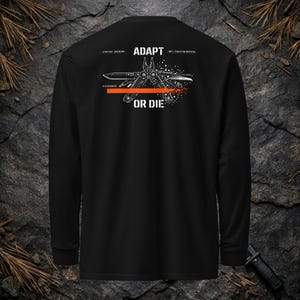 May include: A black long-sleeve shirt featuring a white graphic of a multi-tool with pliers and blades. The text ADAPT OR DIE is printed in white above and below the tool, accompanied by a horizontal orange line and small binary code accents.