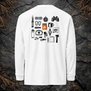 Può includere: Una maglietta bianca a maniche lunghe con una stampa grafica sul retro. Il design è composto da icone nere che rappresentano attrezzatura da campeggio, tra cui un coltello, corda, bussola, binocolo, torcia, kit di pronto soccorso e un'ascia.