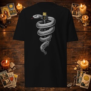 Op de afbeelding: Een zwart t-shirt met een grafische print van een witte slang die om een gouden USB-connector is gewikkeld. De slang heeft een rode tong en de staart gaat over in een gepixeld patroon. Het ontwerp is gecentreerd op de voorkant van het kledingstuk.