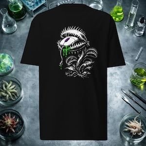 May include: A black t-shirt featuring a white graphic illustration of a Venus flytrap plant with jagged teeth, consuming a computer mouse with a purple scroll wheel and dripping bright green slime from its mouth.