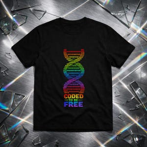 Puede incluir: Camiseta negra con un diseño de hélice de ADN arcoíris compuesto por código binario. Las palabras "CODED TO BE FREE" están impresas debajo en un degradado arcoíris. La camiseta se muestra sobre una superficie reflectante.