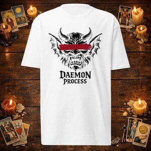 Op de afbeelding: Een wit t-shirt met korte mouwen voorzien van een zwarte afbeelding van een gehoornd demonengezicht met een rode horizontale penseelstreek over de ogen. Onder de afbeelding staat de tekst DAEMON PROCESS in een vetgedrukt, zwart schreeflettertype.