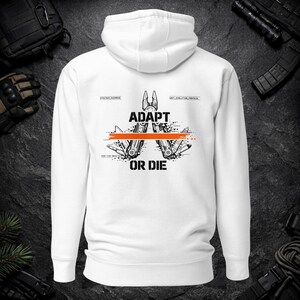 May include: A white hooded sweatshirt featuring a black graphic on the back. The design displays a multi-tool illustration with an orange horizontal line, the text ADAPT OR DIE, and smaller technical labels including STRATEGY_OVERRIDE.