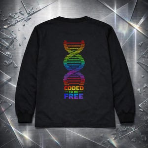 Puede incluir: Camiseta negra de manga larga con un diseño de cadena de ADN arcoíris hecho de código binario. El texto "CODED TO BE FREE" está debajo, también en colores del arcoíris.