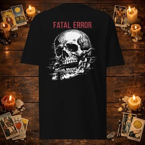 Puede incluir: Una camiseta negra de manga corta con un gráfico blanco de un cráneo humano con un efecto de distorsión digital. Sobre el cráneo, el texto FATAL ERROR está impreso en rojo. El diseño está centrado en el pecho de la prenda con cuello redondo.