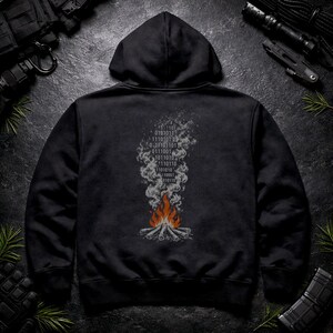 Puede incluir: Una sudadera con capucha negra que presenta un gráfico en la parte posterior. El diseño muestra una fogata con llamas naranjas y rojas que surgen de leños, transformándose en humo gris que forma una columna de código binario de unos y ceros.