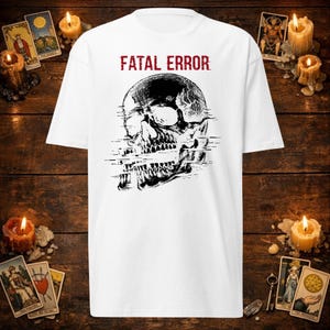 Puede incluir: Una camiseta blanca de manga corta con un gráfico negro de un cráneo humano con un efecto de fallo digital en la mandíbula. El texto "FATAL ERROR" está impreso en letras mayúsculas rojas sobre la ilustración del cráneo en la parte frontal.