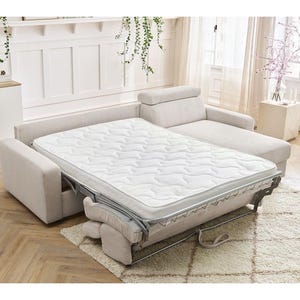 May include: A beige sectional sofa with a pull-out bed. The bed has a white mattress with a quilted pattern. The sofa is on a light-coloured rug. The room has white walls and a wooden floor.