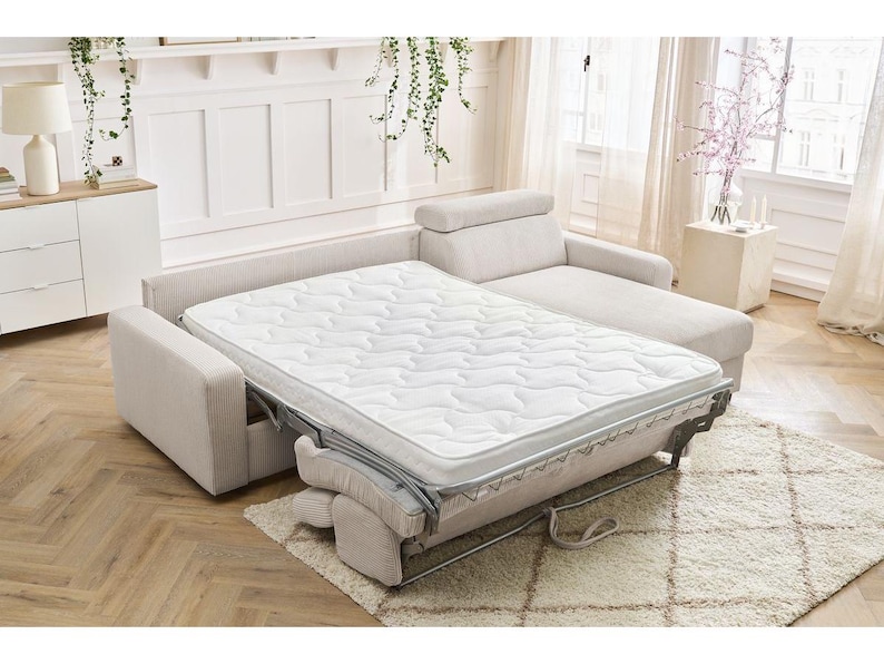 May include: A beige sectional sofa with a pull-out bed. The bed has a white mattress with a quilted pattern. The sofa is on a light-coloured rug. The room has white walls and a wooden floor.