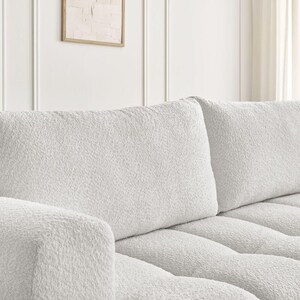 Op de afbeelding: Close-up van een deel van een witte, getextureerde bank. De bank heeft een zachte, pluche uitstraling met zichtbare tufting op de zitkussens. De rugkussens zijn rechthoekig en hebben ook een getextureerd oppervlak.