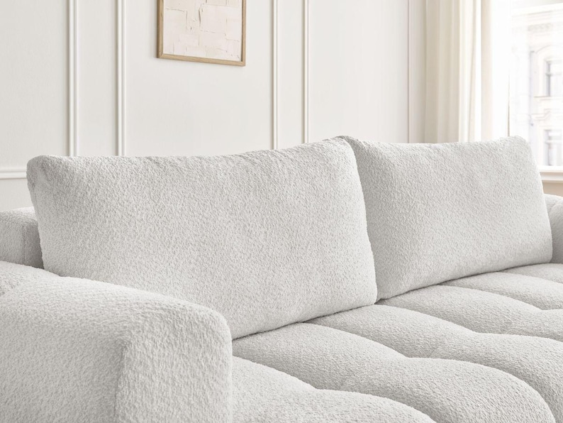 Op de afbeelding: Close-up van een deel van een witte, getextureerde bank. De bank heeft een zachte, pluche uitstraling met zichtbare tufting op de zitkussens. De rugkussens zijn rechthoekig en hebben ook een getextureerd oppervlak.