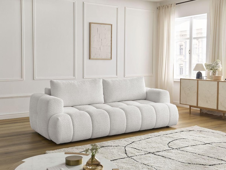 Op de afbeelding: Een grote, witte, moderne bank met een getextureerd, gecapitonneerd ontwerp. De bank heeft twee grote rugkussens en afgeronde armleuningen. De kamer heeft witte muren, een lichtgekleurd tapijt en een ingelijst kunstwerk. De bank is ongeveer 220 cm lang.