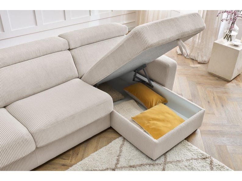 May include: Beige corduroy sectional sofa with an open storage compartment containing two yellow cushions. The sofa sits on a patterned rug. A small side table with a vase of flowers is also visible.