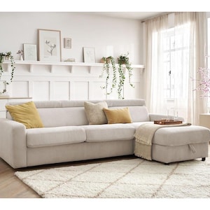 May include: A beige corduroy sectional sofa with a matching ottoman. The sofa has three seat cushions and several decorative cushions in shades of yellow and beige. A wooden tray with glasses sits on the ottoman, and a cream-coloured throw blanket is draped over it.