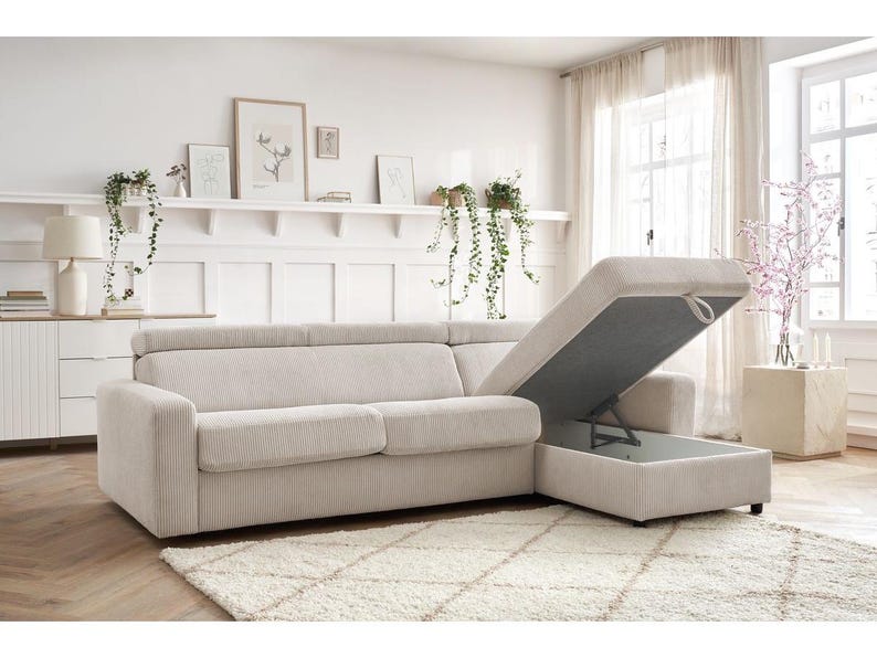 May include: A beige corduroy sectional sofa with a storage compartment. The sofa is on a patterned rug. The storage compartment is open, revealing the interior. The room has a neutral colour palette with decorative plants and artwork.