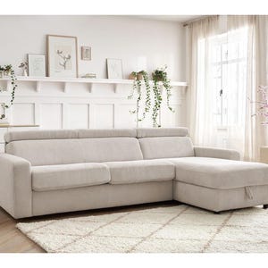 May include: A light beige corduroy sectional sofa with a chaise lounge. The sofa sits on a cream-coloured rug. The room features white walls, a white cabinet, and a window with sheer curtains. The sofa is a comfortable addition to any living room.