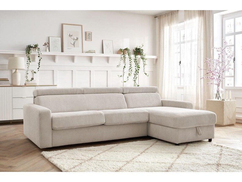 May include: A light beige corduroy sectional sofa with a chaise lounge. The sofa sits on a cream-coloured rug. The room features white walls, a white cabinet, and a window with sheer curtains. The sofa is a comfortable addition to any living room.
