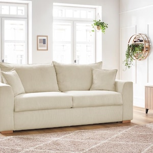 Peut inclure: Un canap&eacute; en velours c&ocirc;tel&eacute; cr&egrave;me avec deux coussins d'assise et deux coussins de dossier. Le canap&eacute; a deux coussins d&eacute;coratifs assortis et des pieds en bois. La pi&egrave;ce comprend de grandes fen&ecirc;tres, un tapis et des plantes d&eacute;coratives.