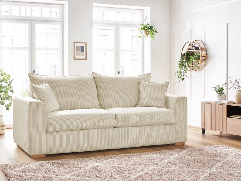 Peut inclure: Un canap&eacute; en velours c&ocirc;tel&eacute; cr&egrave;me avec deux coussins d'assise et deux coussins de dossier. Le canap&eacute; a deux coussins d&eacute;coratifs assortis et des pieds en bois. La pi&egrave;ce comprend de grandes fen&ecirc;tres, un tapis et des plantes d&eacute;coratives.