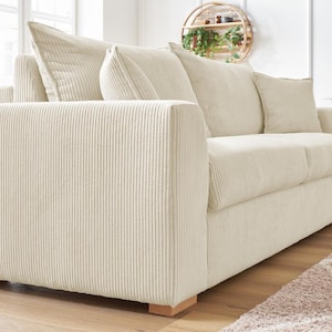 Peut inclure: Un canap&eacute; en velours c&ocirc;tel&eacute; cr&egrave;me avec trois coussins d'assise et plusieurs coussins de dossier. Le canap&eacute; a une forme carr&eacute;e et des pieds en bois. Le tissu en velours c&ocirc;tel&eacute; a une texture c&ocirc;tel&eacute;e verticale. Le canap&eacute; est dans une pi&egrave;ce lumineuse.