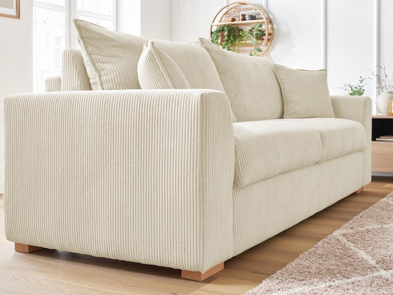 Peut inclure: Un canap&eacute; en velours c&ocirc;tel&eacute; cr&egrave;me avec trois coussins d'assise et plusieurs coussins de dossier. Le canap&eacute; a une forme carr&eacute;e et des pieds en bois. Le tissu en velours c&ocirc;tel&eacute; a une texture c&ocirc;tel&eacute;e verticale. Le canap&eacute; est dans une pi&egrave;ce lumineuse.