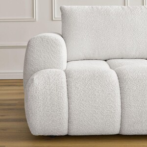 Op de afbeelding: Een gedeelte van een witte, getextureerde bank met een afgeronde armleuning en een rechthoekige rugleuning. De bank heeft een zachte boucl&eacute; stof en een modern design. De achtergrond toont een witte muur met lijstwerk en een houten vloer.