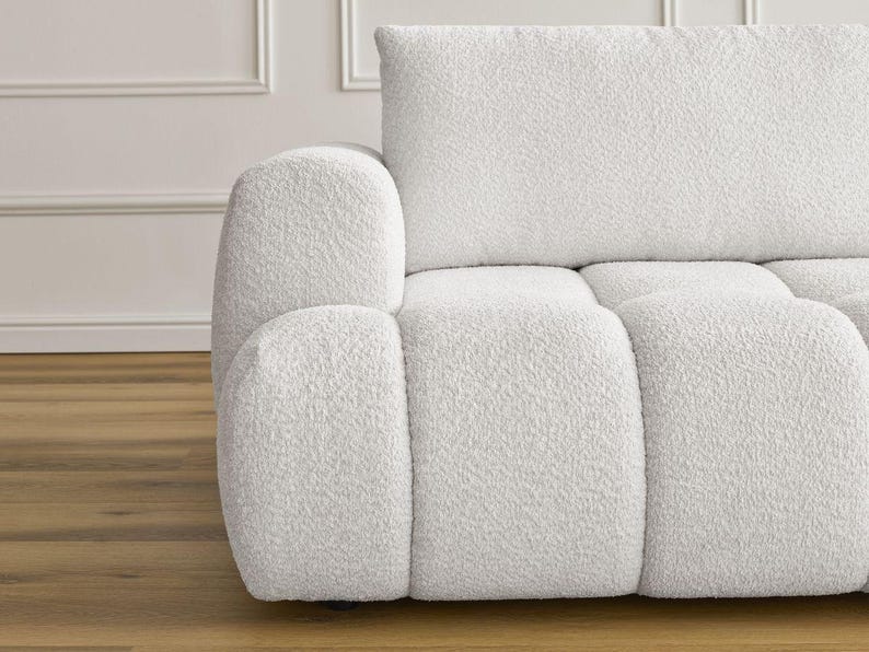 Op de afbeelding: Een gedeelte van een witte, getextureerde bank met een afgeronde armleuning en een rechthoekige rugleuning. De bank heeft een zachte boucl&eacute; stof en een modern design. De achtergrond toont een witte muur met lijstwerk en een houten vloer.