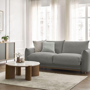 Puede incluir: Salón moderno con sofá gris, cojines decorativos, mesa de centro redonda con tapa blanca y patas marrones, y aparador gris. Una alfombra beige y cortinas transparentes completan la paleta de colores neutros.