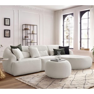 May include: A contemporary living room with a large, cream-coloured sectional sofa and cushions. A matching ottoman and a patterned rug are in front of the sofa. A gold side table and a black metal shelving unit are also present.