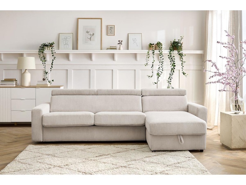 May include: A cream corduroy sectional sofa with a chaise lounge on the right. The sofa is on a cream rug. The background features a white wall, artwork, and hanging plants. The sofa is approximately 250 cm long.