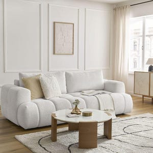 Op de afbeelding: Een moderne woonkamer met een witte boucl&eacute; bank, een ronde salontafel en een patroon tapijt. De kamer heeft een neutraal kleurenpalet met een ingelijst kunstwerk en een houten kast. Zacht licht komt door het raam.