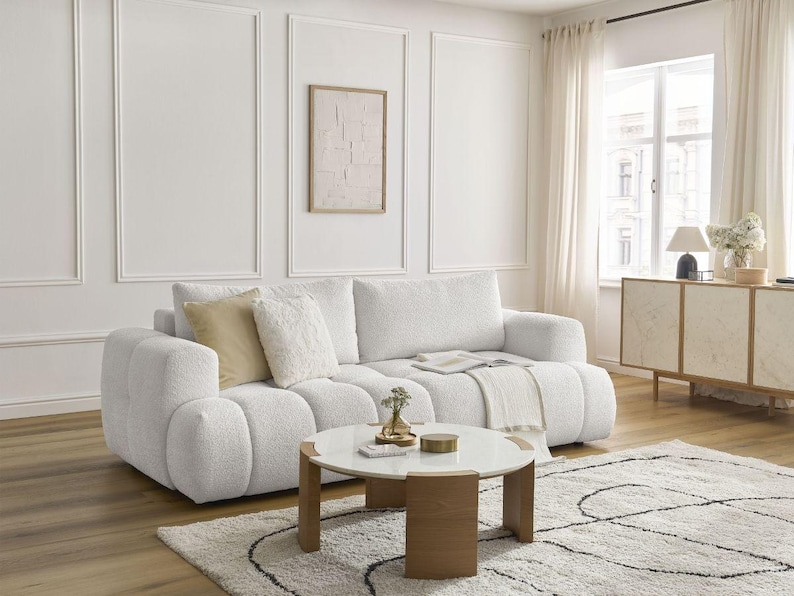 Op de afbeelding: Een moderne woonkamer met een witte boucl&eacute; bank, een ronde salontafel en een patroon tapijt. De kamer heeft een neutraal kleurenpalet met een ingelijst kunstwerk en een houten kast. Zacht licht komt door het raam.