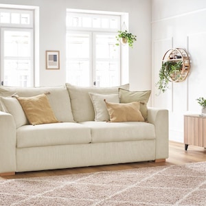 Peut inclure: Un canap&eacute; en velours c&ocirc;tel&eacute; cr&egrave;me avec plusieurs coussins beiges et bruns. Le canap&eacute; est pos&eacute; sur un tapis marron clair. La pi&egrave;ce est orn&eacute;e de grandes fen&ecirc;tres et de plantes d&eacute;coratives.