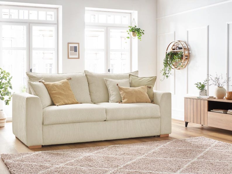 Peut inclure: Un canap&eacute; en velours c&ocirc;tel&eacute; cr&egrave;me avec plusieurs coussins beiges et bruns. Le canap&eacute; est pos&eacute; sur un tapis marron clair. La pi&egrave;ce est orn&eacute;e de grandes fen&ecirc;tres et de plantes d&eacute;coratives.