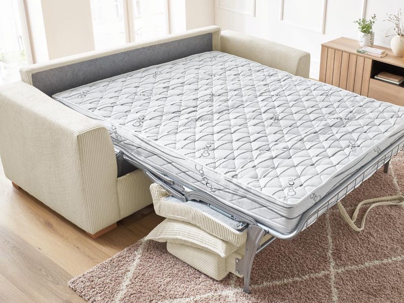 Peut inclure: Un canap&eacute;-lit beige avec un matelas d&eacute;pliable. Le matelas est blanc avec un motif matelass&eacute; et un cadre en m&eacute;tal argent&eacute;. Le canap&eacute; a un dossier gris et des accoudoirs en velours c&ocirc;tel&eacute; beige. Des oreillers sont empil&eacute;s &agrave; la base du lit.