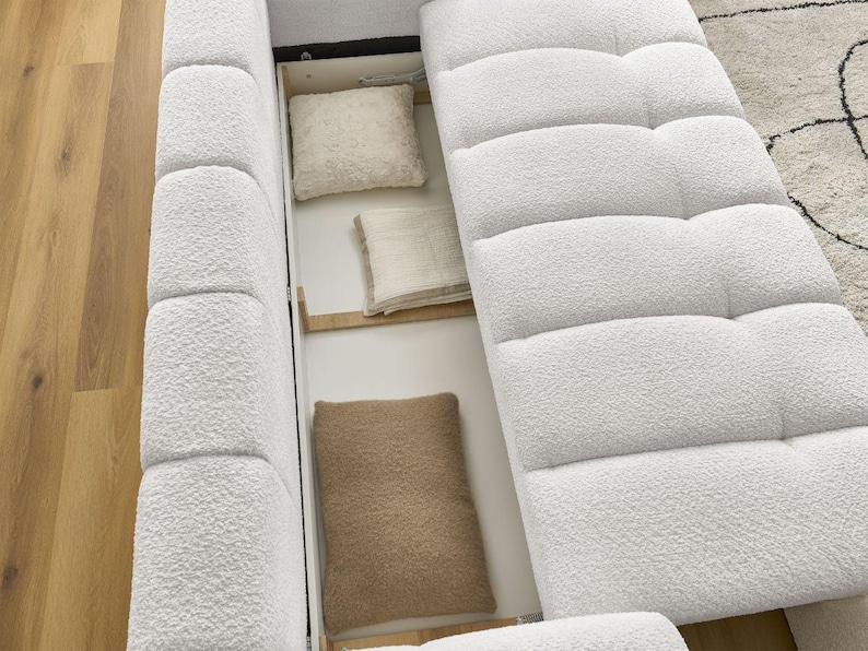 Op de afbeelding: Een witte, getextureerde bank met een opklapbaar gedeelte dat opbergruimte onthult. Binnenin bevinden zich drie kussens in cr&egrave;me- en bruintinten. De bank staat op een lichte houten vloer, met een patroonkleed zichtbaar op de achtergrond.