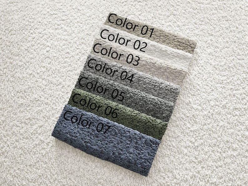 Op de afbeelding: Een kleurenpalet van zeven stofstalen, elk met een andere tint. De kleuren vari&euml;ren van lichtbeige tot donkerblauw, met de tekst "Color 01" tot "Color 07" op elk staal. De stalen zijn diagonaal gerangschikt op een wit, getextureerd oppervlak.