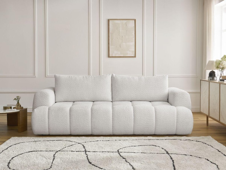 Op de afbeelding: Een witte, getextureerde bank met twee grote rugkussens en een laag profiel. De bank heeft een afgeronde vorm en staat op een lichtgekleurd tapijt met een zwart abstract ontwerp. De kamer heeft een neutraal kleurenpalet.