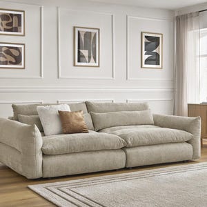Op de afbeelding: Een grote, beige hoekbank met meerdere zachte kussens en kussens. De bank staat in een woonkamer, met ingelijste abstracte kunst aan de muur en een tapijt in neutrale tinten. Een houten kast en transparante gordijnen zijn ook zichtbaar.